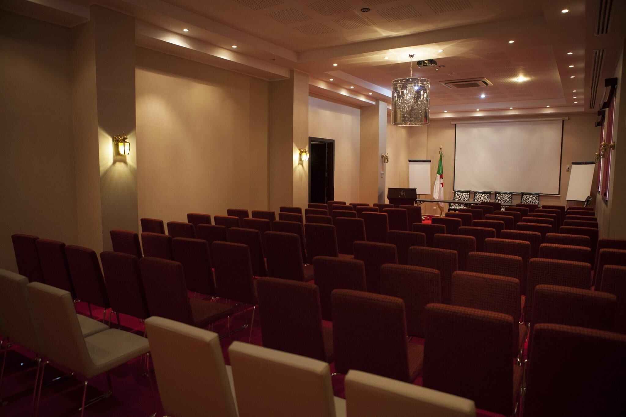 Un salón de conferencias con filas de sillas, un atril, una pantalla de proyector y una bandera.