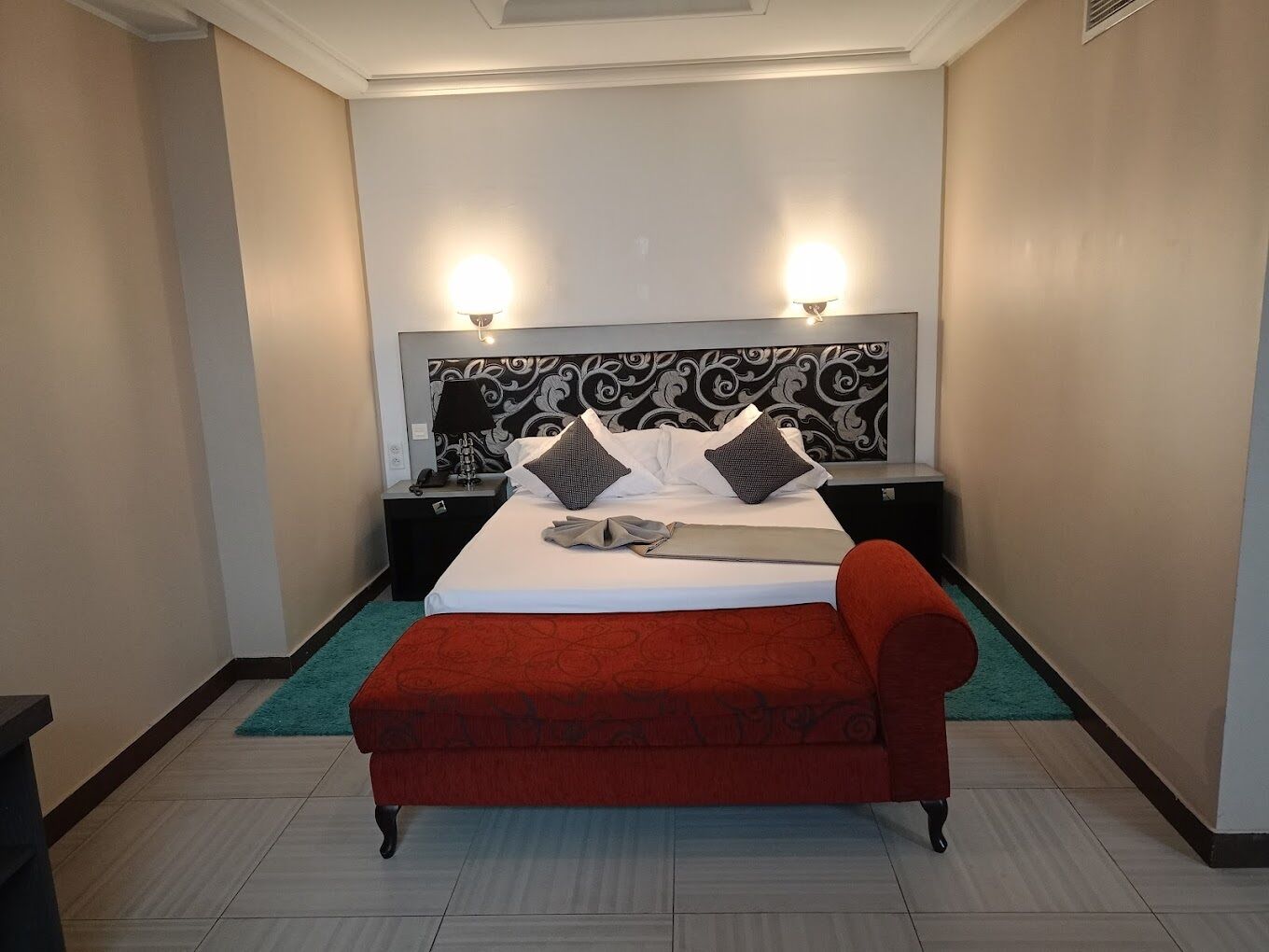 Una habitación de hotel con una cama, dos mesitas de noche, un banco rojo y un cabecero estampado.