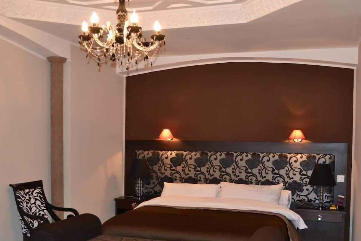 A bedroom with a large bed, a chandelier, patterned wallpaper, and a nightstand with a lamp.