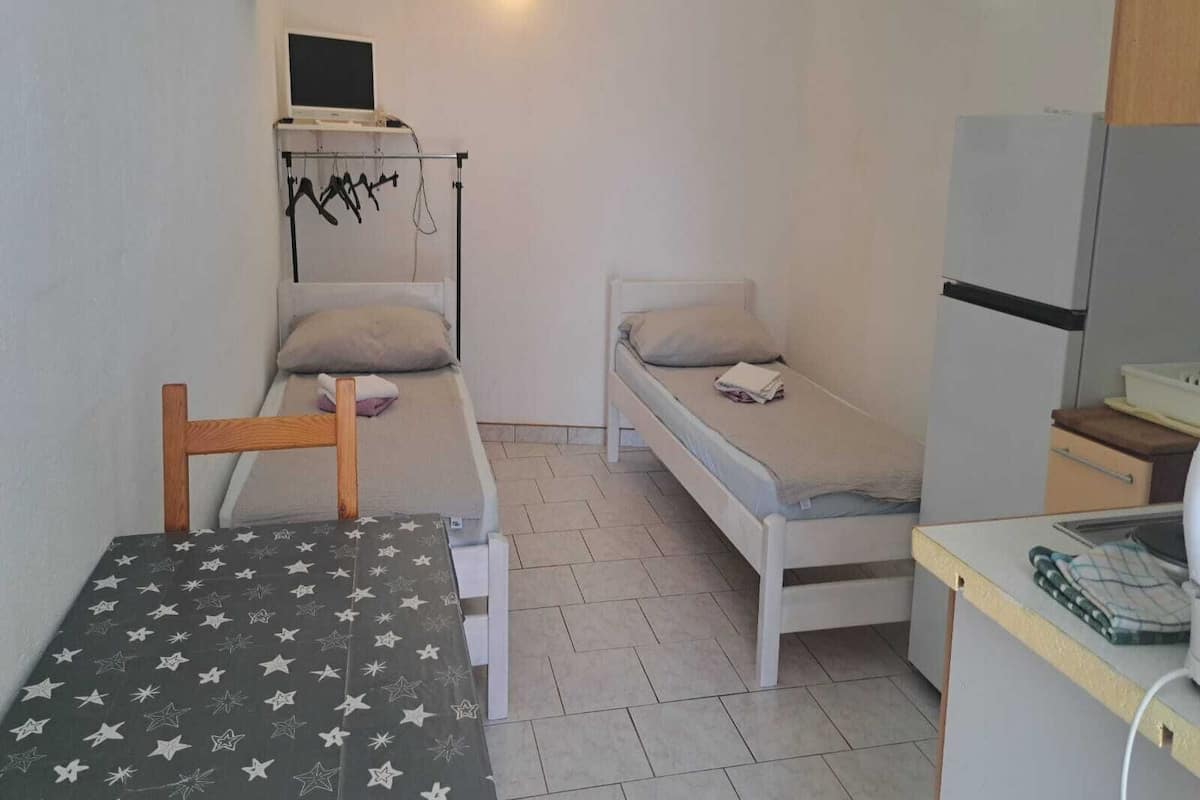 A compact room with two single beds, a small table, a kitchenette, and a wall-mounted TV.
