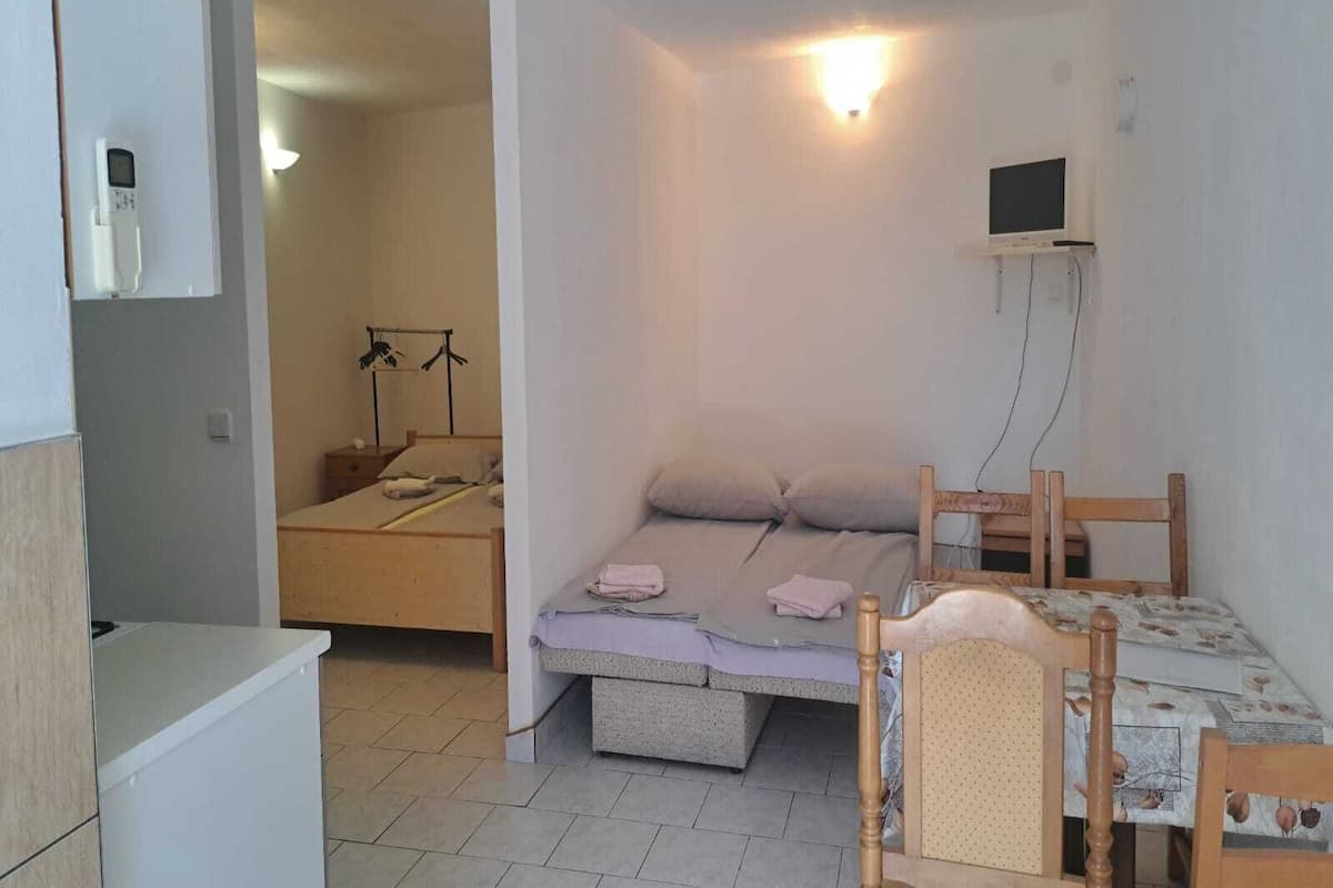 A small, well-lit room with a bed, a desk, and a chair. There is a television mounted on the wall and a shelf above the bed.