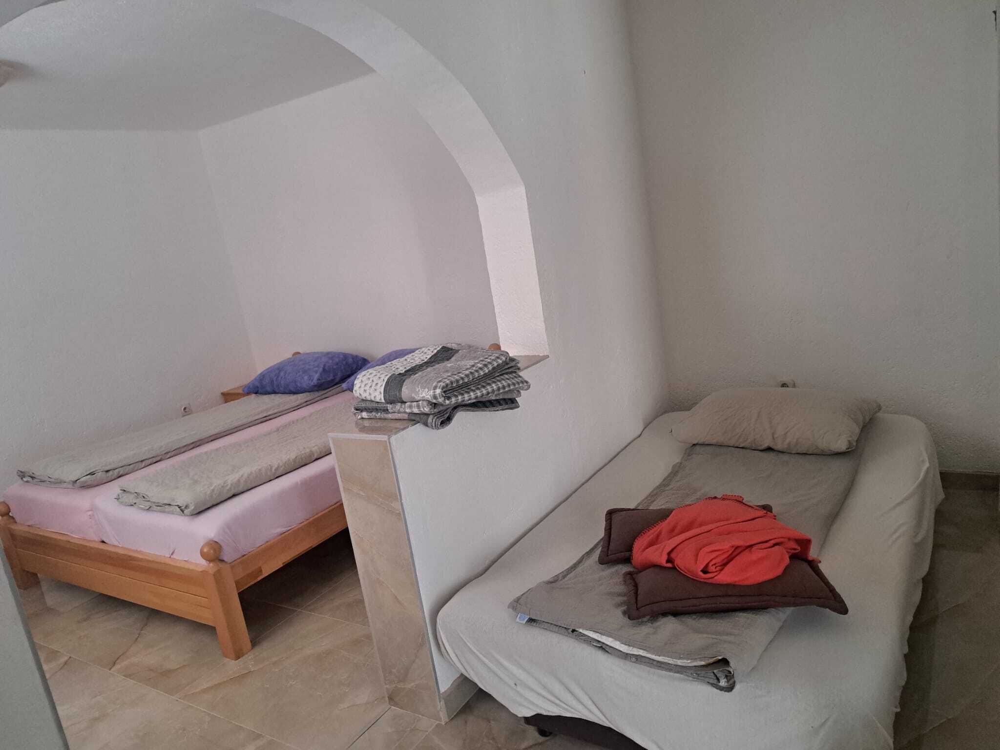 A small, single-room accommodation with two beds, one with a wooden frame and the other with a metal frame. There is a small table with a folded blanket and a pillow on it.