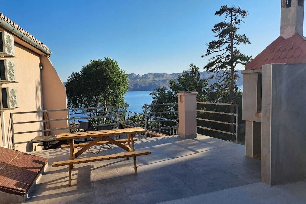 An outdoor terrace with a wooden table and chairs, a view of the sea, and a mountain range in the background.