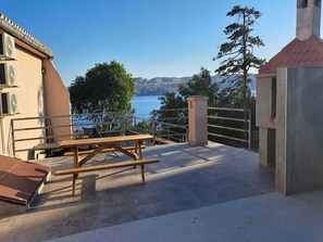 Una terraza exterior con mesa y sillas de madera, vista al mar y una cordillera en el fondo.