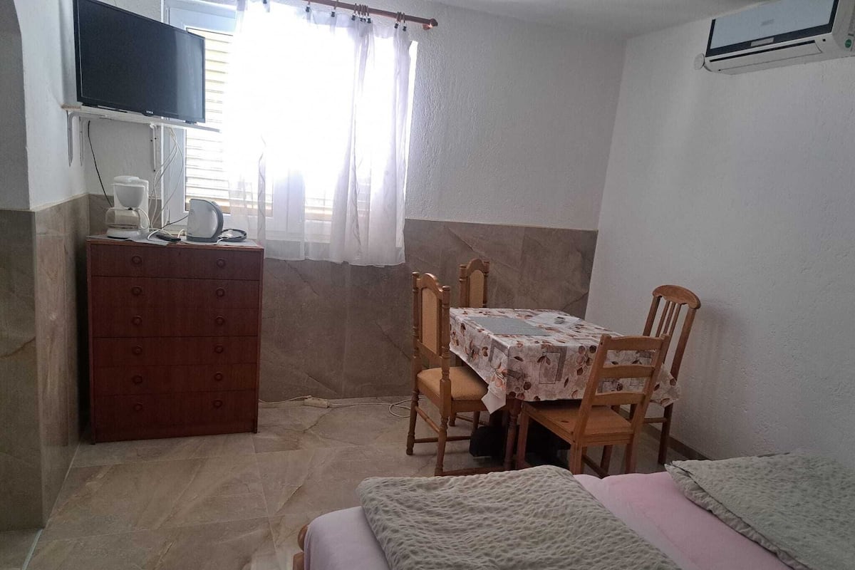 A small, well-lit room with a bed, a table with chairs, a wooden dresser, and a television mounted on the wall.