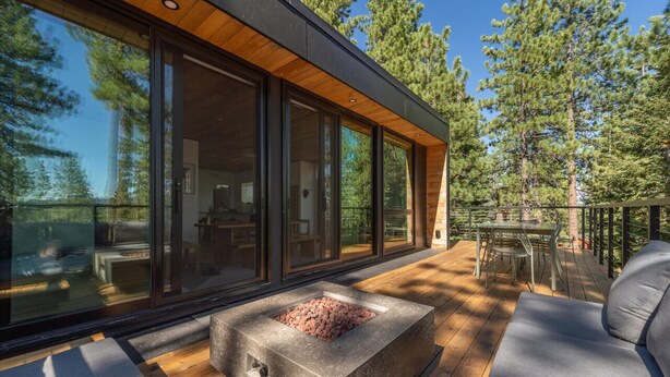 A modern house with large glass windows, a stone fire pit, and a wooden deck.