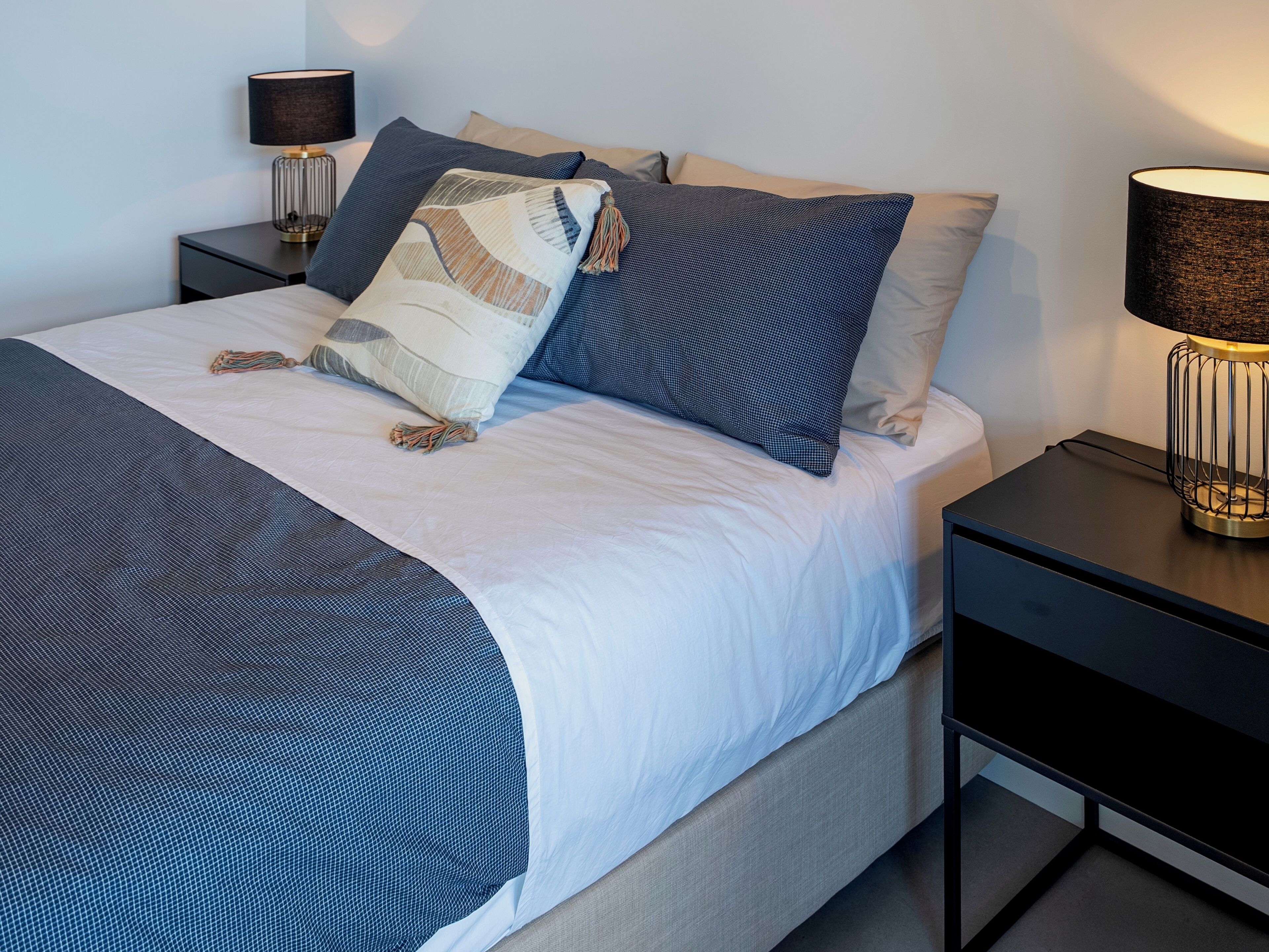 Una cama bien hecha con colcha azul y blanca, dos mesitas de noche con lámparas, y un pequeño aparador junto a la cama.