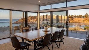 A modern dining area with a large table and chairs, large windows offering a view of the sea and hills. - Cove Landing Ocean View Apt-01 Karatta Sleeps 4 (Penneshaw)
