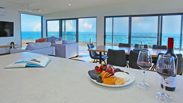 A modern living room with a large window overlooking the sea, a dining area with a table set for two, and a sofa with a book on it. - Cove Landing Ocean View Apt-01 Karatta Sleeps 4 (Penneshaw)