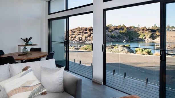 A modern living room with a large sliding glass door leading to a balcony with a scenic view of a lake and hills. - Cove Landing Ocean View Apt-02 James Comrie Sleeps 4 (Penneshaw)