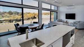 A modern kitchen with a large island, a dining area, and a living space with a large window offering a view of the outdoors. - Cove Landing Ocean View Apt-02 James Comrie Sleeps 4 (Penneshaw)