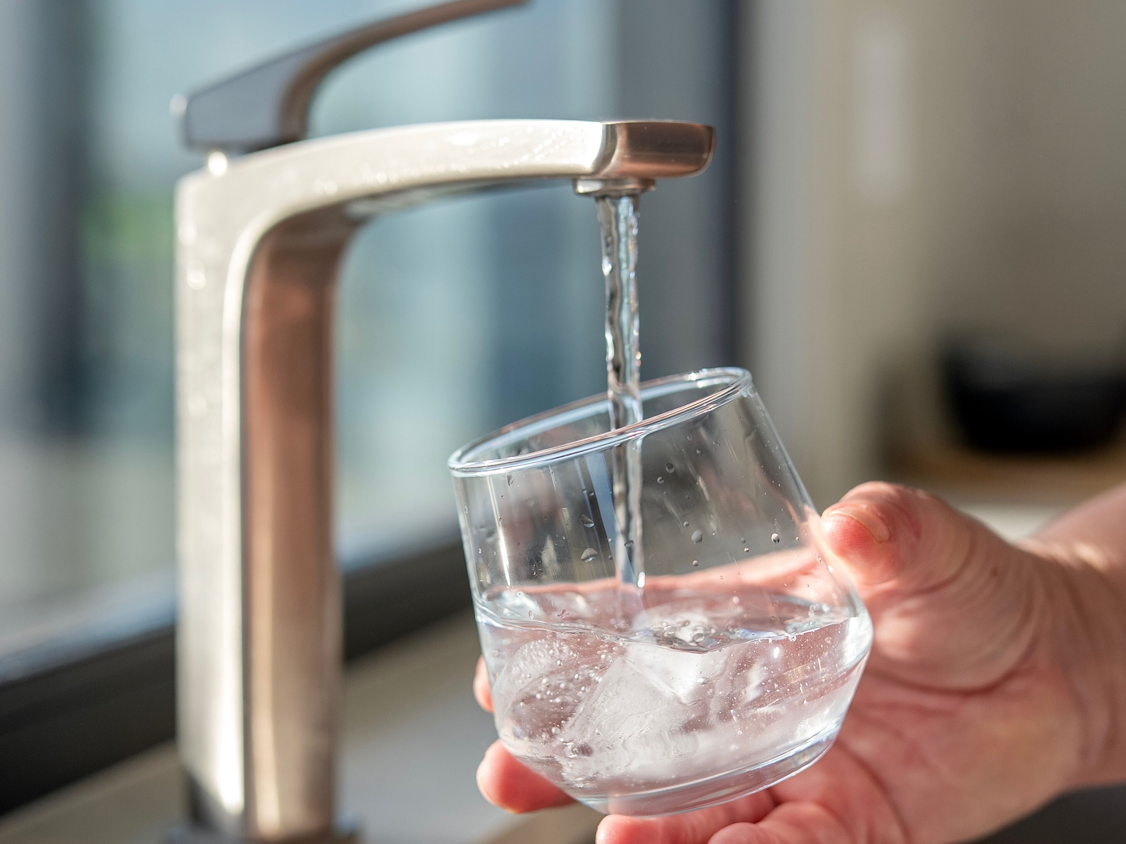 A hand holding a glass with water and ice under a running tap.