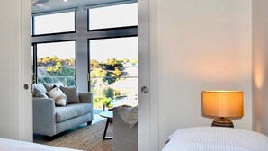 A modern hotel room with a large window, a sofa, a small table, and a lamp. - Cove Landing Ocean View Apt-02 James Comrie Sleeps 4 (Penneshaw)