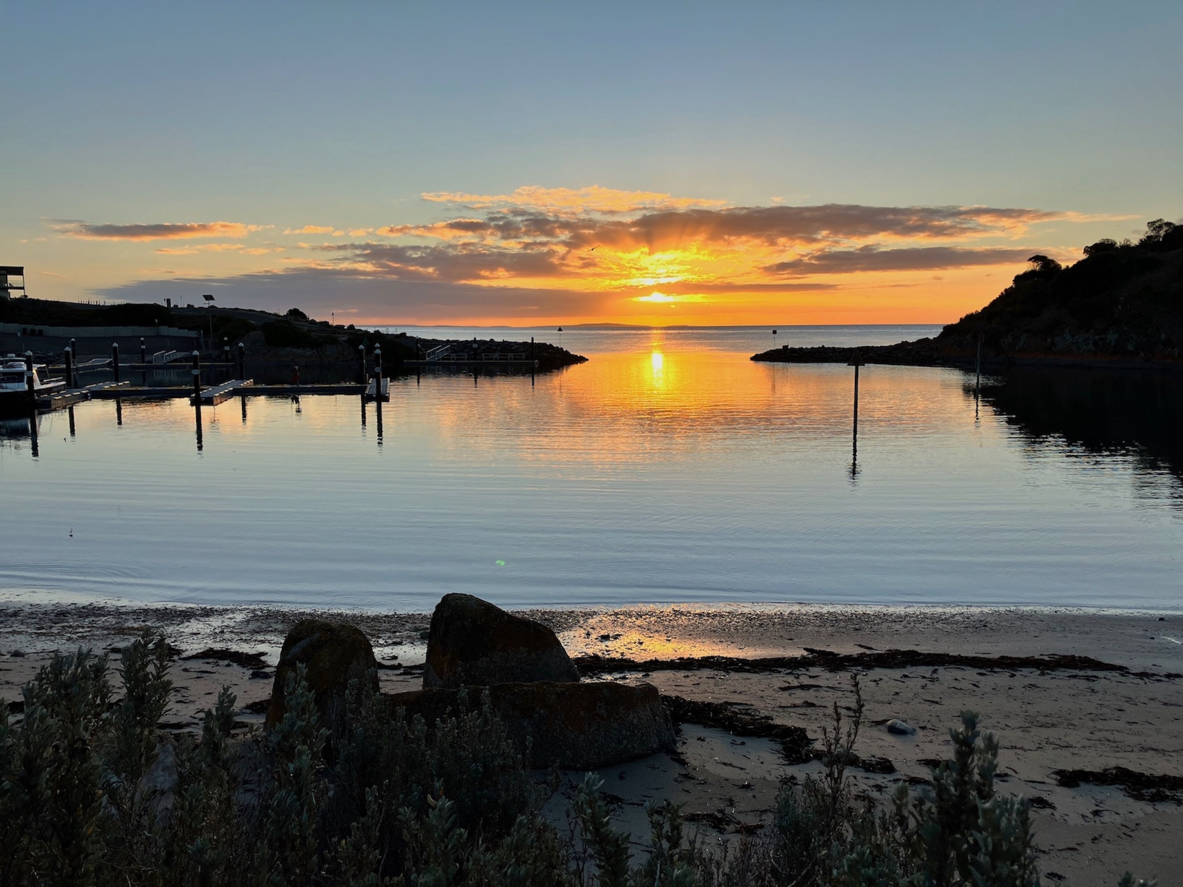 A sunset over a calm body of water with a pier and rocky shoreline.