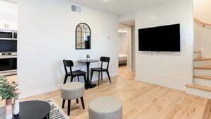 A modern living room with a dining area, a flat-screen TV mounted on the wall, and a staircase leading to another level. - Lofty Urban Retreat (Bridgeview)
