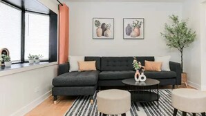 A modern living room with a dark grey sectional sofa, two framed floral artworks on the wall, a round coffee table with a vase and a pair of white candles, and a potted plant to the right. - Lofty Urban Retreat (Bridgeview)