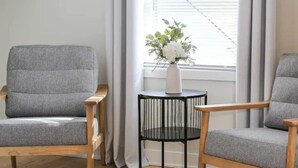 A room with two grey upholstered chairs, a small black table with a white vase of flowers, and a window with white curtains. - Lofty Urban Retreat (Bridgeview)