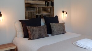 A neatly made bed with a white duvet and two patterned pillows, a wooden nightstand with a power outlet, and a wall-mounted abstract art piece above the bed. - Quinta da Janela Azul - Limão (Tavira)