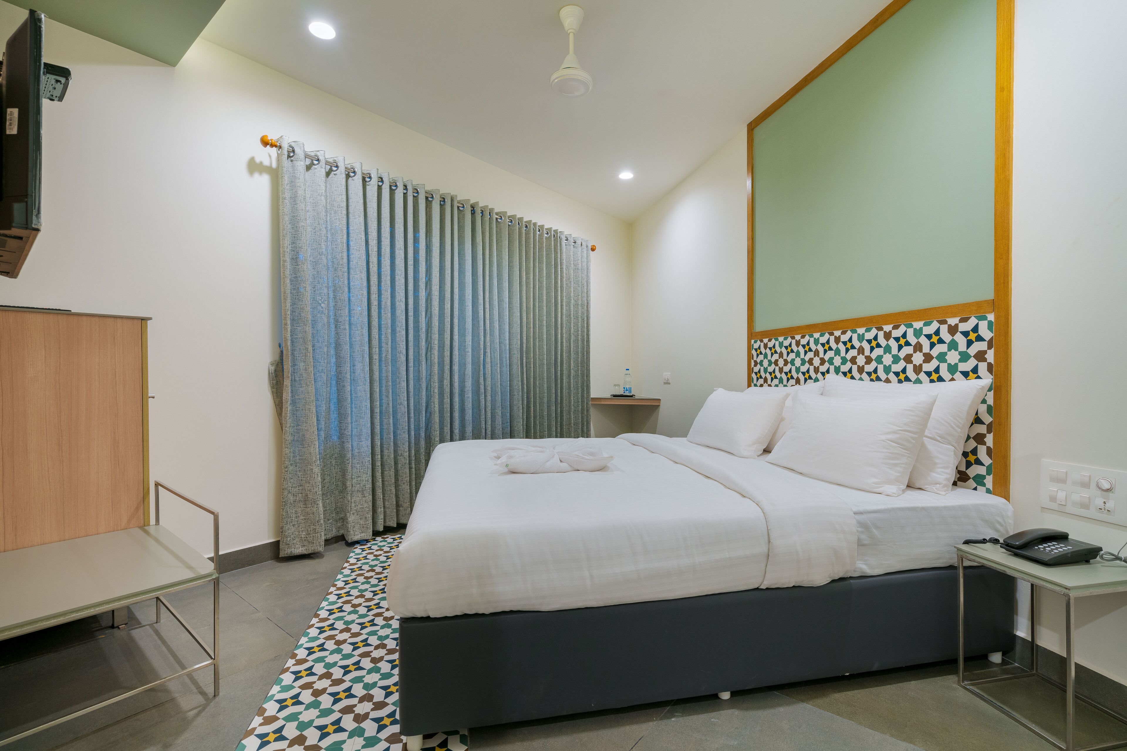 A modern hotel room with a large bed, a bedside table with a phone, a small table with a lamp, and a curtain.