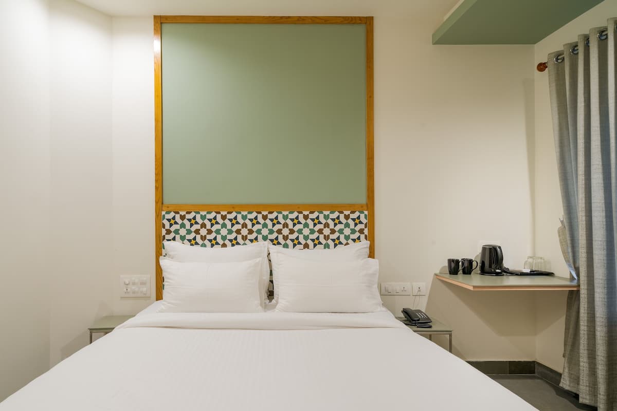 A hotel room with a bed, a bedside table with a phone, a small shelf with a kettle and mugs, and a patterned headboard.