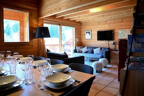 A wooden cabin interior with a dining table set for four, a sofa, a TV, and a fireplace.