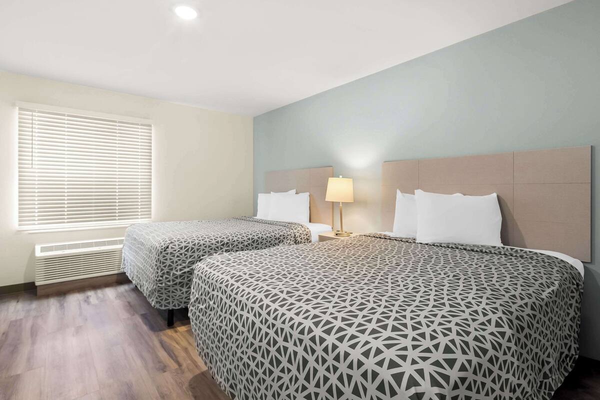 A hotel room with two beds, a bedside table with a lamp, a window with blinds, and a wall-mounted heater.