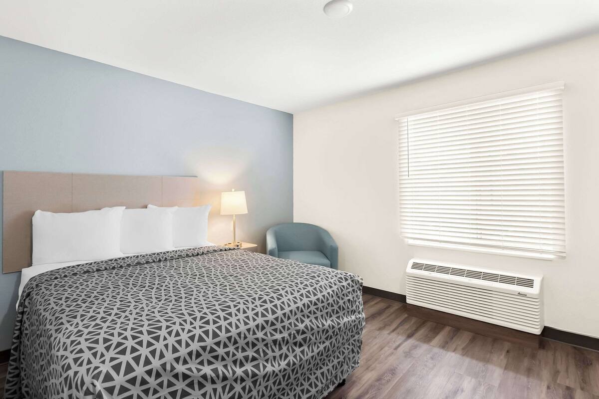 A hotel room with a bed, a chair, a lamp, and a window with blinds.