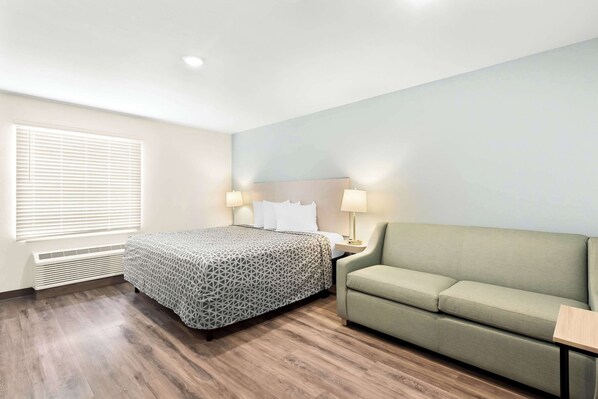 A hotel room with a bed, a green sofa, a nightstand with lamps, and a window with blinds. - Woodspring Suites Savannah West Chatham Parkway (Savannah)
