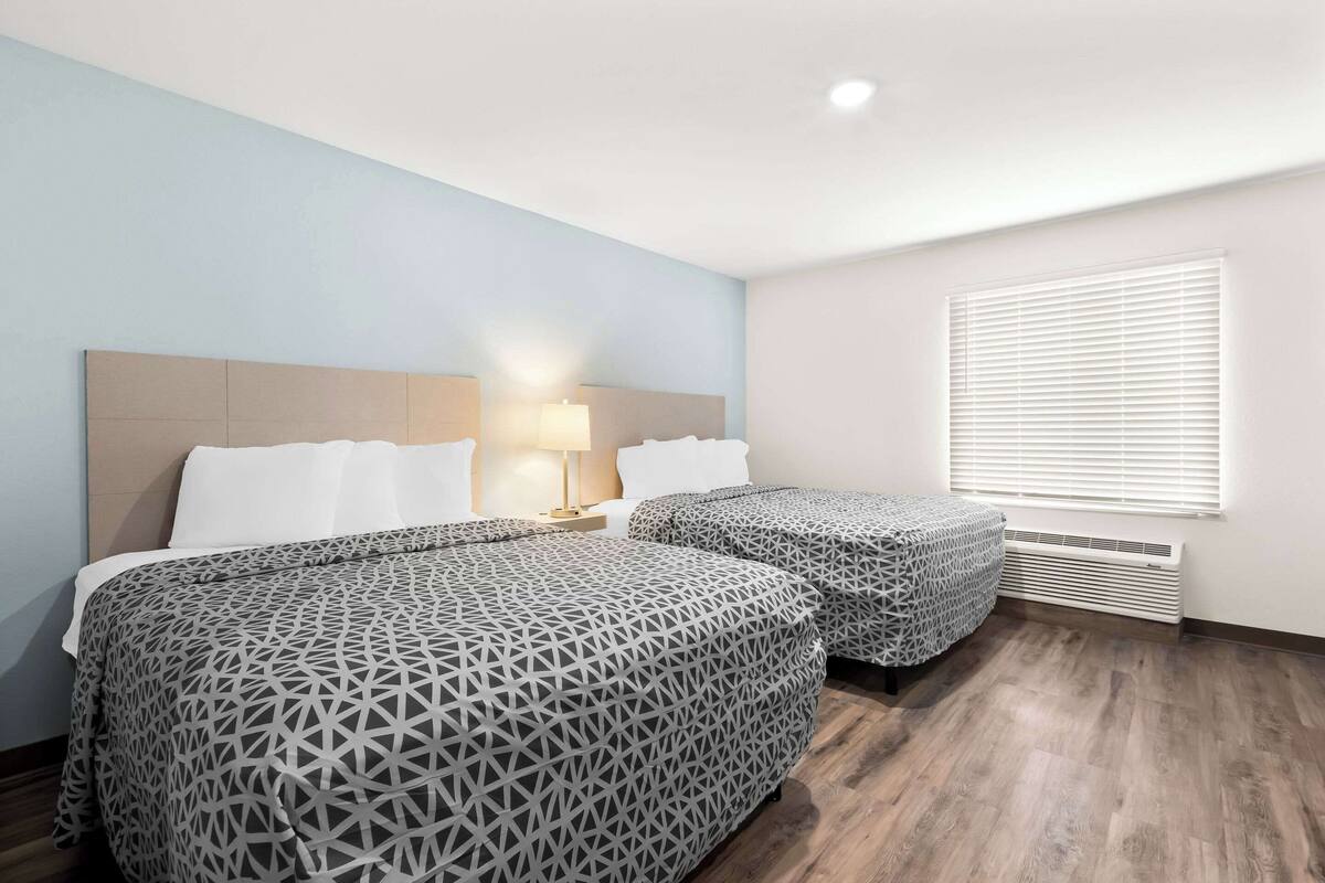 A hotel room with two beds, a nightstand, a lamp, and a window with blinds.