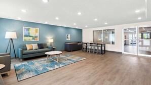 A modern office space with a blue accent wall, a sofa, a dining area, and a large window. - Woodspring Suites Savannah West Chatham Parkway (Savannah)