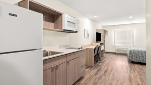 A compact kitchen with a built-in microwave, a refrigerator, and a sink. There is a small dining area with a chair and a TV. The room has wooden flooring and a window with blinds. - Woodspring Suites Savannah West Chatham Parkway (Savannah)