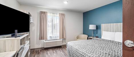 A hotel room with a bed, a desk with a computer, a chair, a window with curtains, and a bedside lamp.