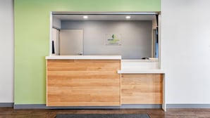 A reception area with a wooden counter, a white wall, and a green accent wall. - WoodSpring Suites Winston-Salem near Hanes Mall (Winston-Salem)