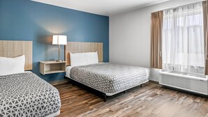 A hotel room with two beds, a nightstand, a lamp, a window with curtains, and an air conditioning unit. - WoodSpring Suites Winston-Salem near Hanes Mall (Winston-Salem)