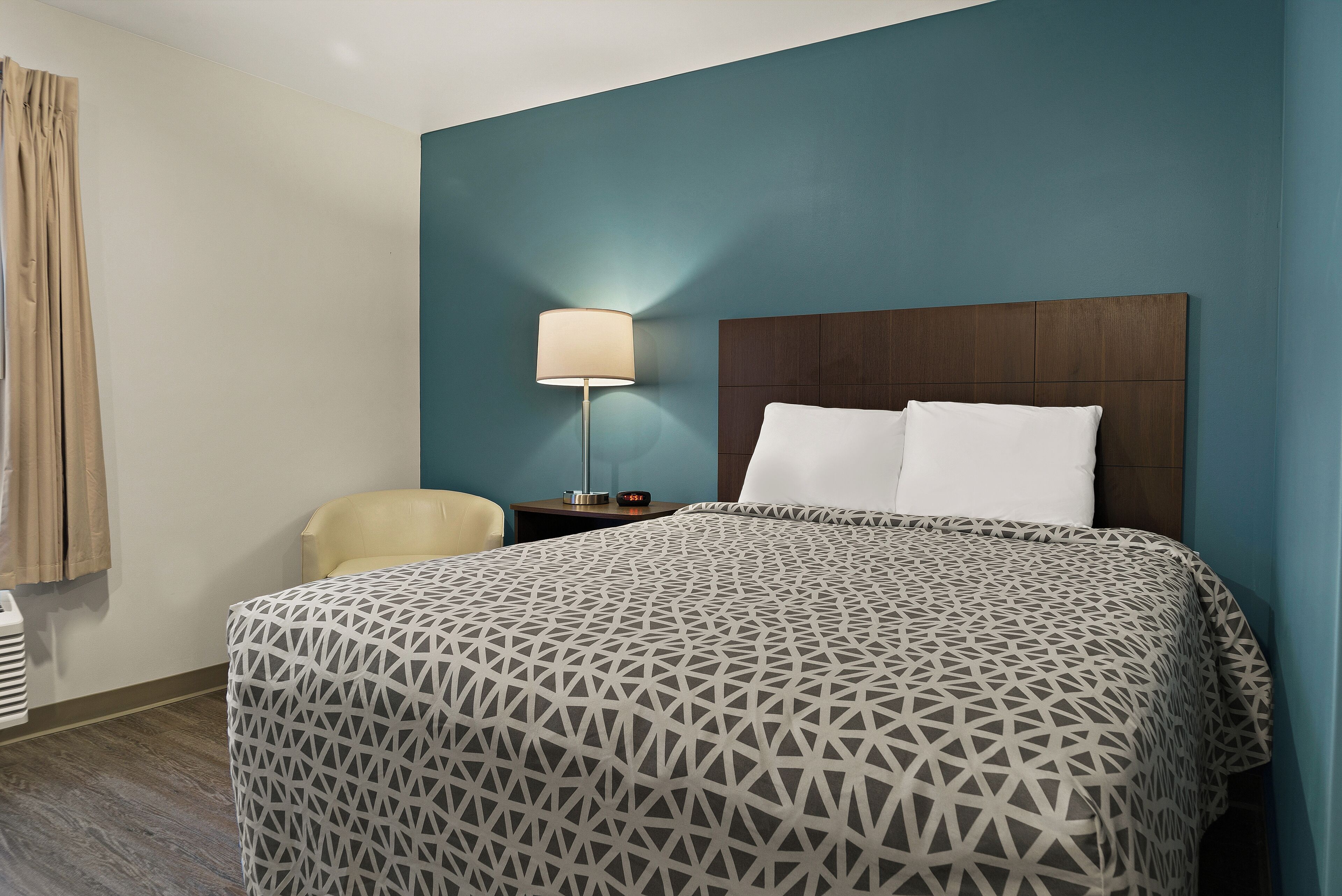 Una habitación de hotel con una colcha estampada, una mesita de noche con lámpara, una silla y una ventana con cortinas.