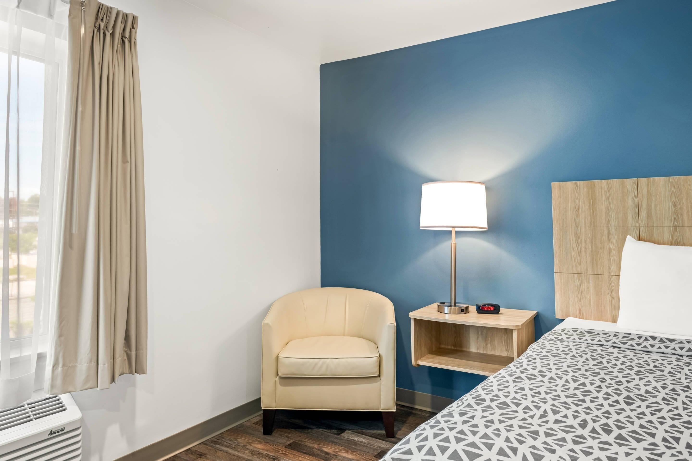 A hotel room with a bed, a chair, a nightstand with a lamp, and a window with curtains.