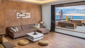 Een moderne woonkamer met een bank, salontafel en poefs. Er is een groot raam met uitzicht op bergen en een waterpartij.