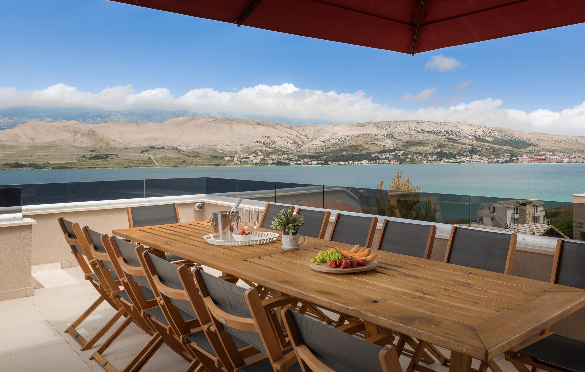 Une table en bois, entourée de chaises, et un parasol rouge, le tout offrant une vue panoramique sur des montagnes et un plan d’eau.