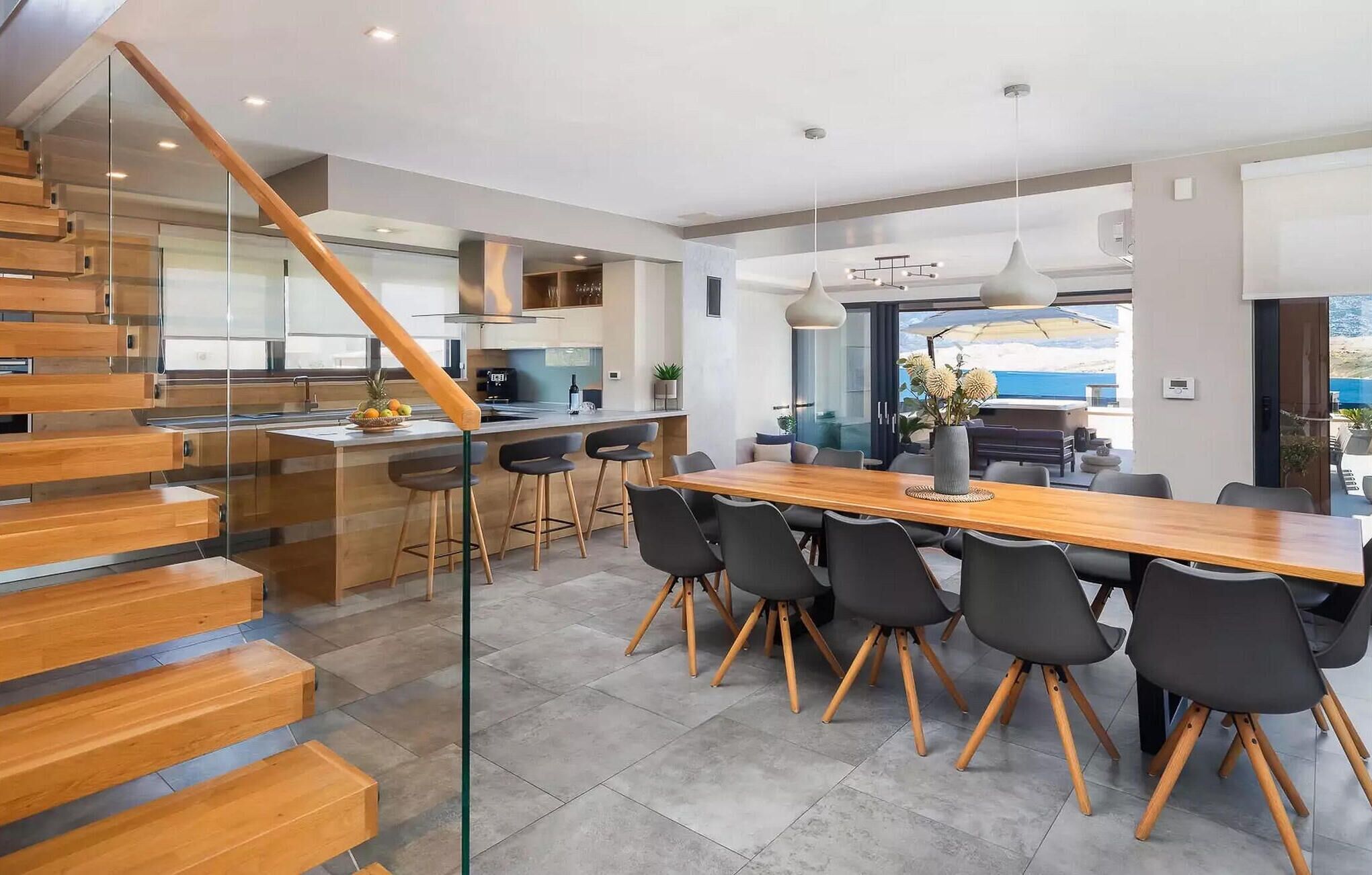 A modern kitchen with a wooden staircase, a dining area with a large table and chairs, and a view of the outdoors.