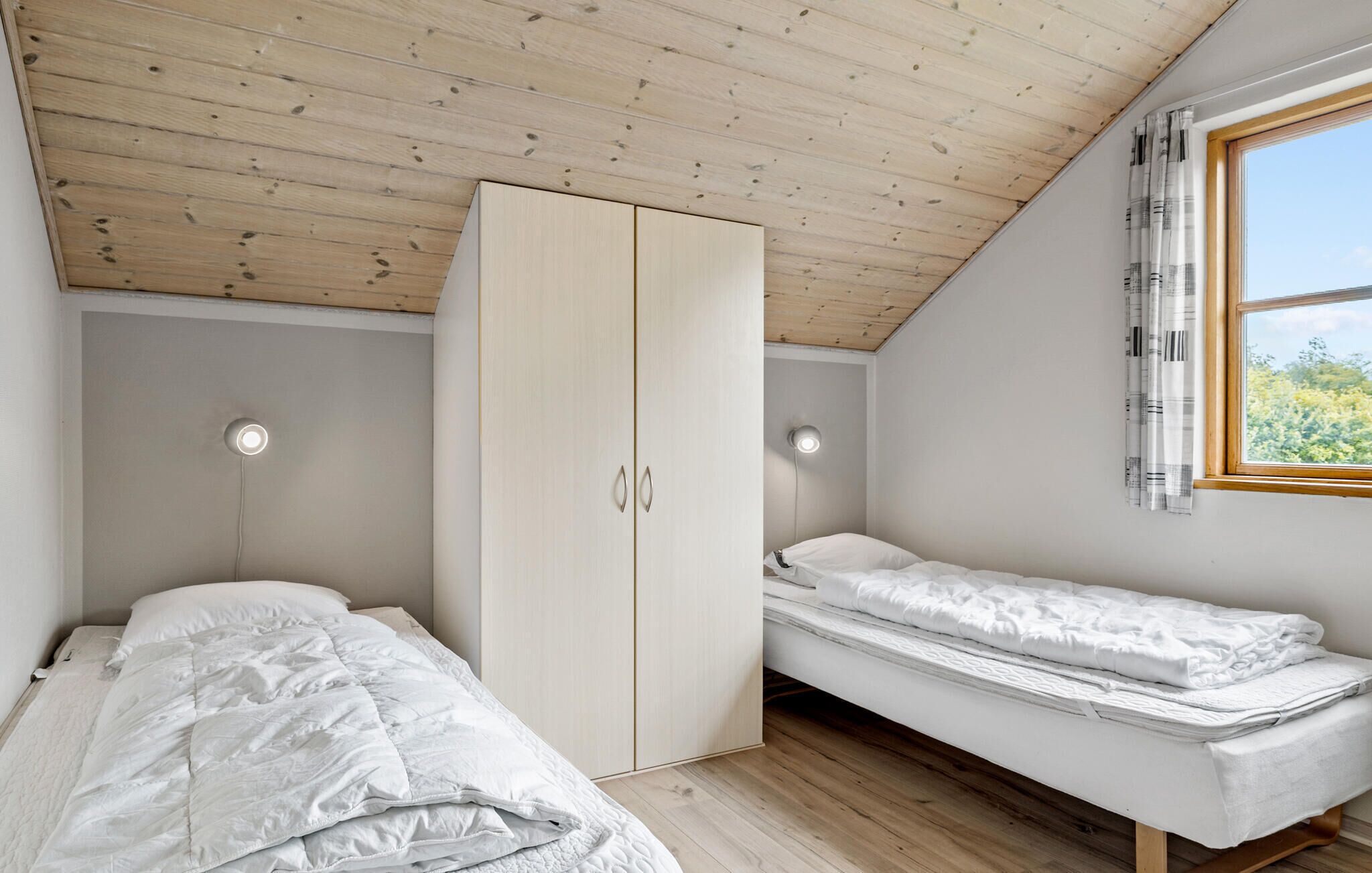 A bedroom with a wooden ceiling, a wardrobe, two beds, and a window with curtains.