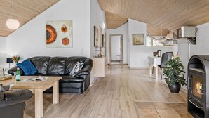 Un vaste espace de vie comprenant un canapé en cuir noir, une table basse en bois et une cheminée. Des tableaux ornent les murs, et on aperçoit une cuisine au fond de la pièce.