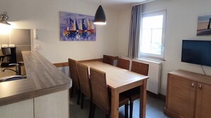 A modern kitchen with a wooden island, dining table with chairs, a painting of sailboats, a hanging pendant light, and a television on the wall. - Appartement Belle Dune 2 Chambres (Fort-Mahon Plage)