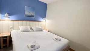 A neatly made bed with white linens and towels, a wooden nightstand, a blue wall with a framed picture, and a white ceiling. - Appartement Belle Dune 2 Chambres (Fort-Mahon Plage)