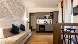 Une cuisine moderne avec des meubles gris, un four à micro-ondes et une cuisinière. Le sol est en bois, il y a un canapé avec des coussins bleus et une grande carte accrochée au mur.