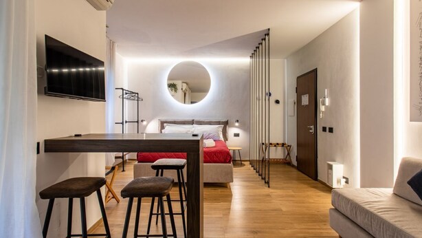 Une chambre d’hôtel moderne avec un plancher en bois, un lit avec une literie rouge, un miroir rond, un téléviseur fixé au mur et un coin bar avec des tabourets.