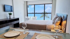 A modern hotel room with a large bed, a TV mounted on the wall, a dining area with two plates and wine glasses, and a view of the ocean through a large window. - Flat Frente Mar R601 (Maceió)