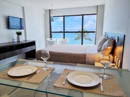 Habitación de hotel moderna con una cama grande, televisor montado en la pared, zona de comedor con dos platos y copas de vino, y vista al océano a través de un ventanal amplio.