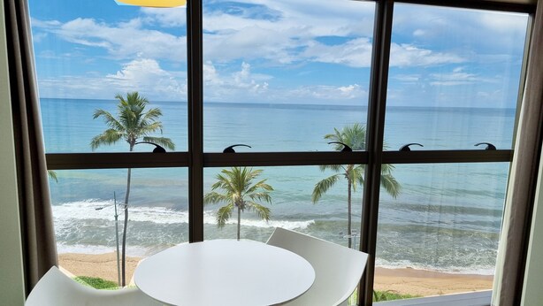A room with a large window overlooking a beach with palm trees and the ocean. There is a round table and two chairs in the room. - Flat Frente Mar R601 (Maceió)