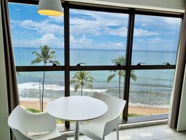 Una habitación con un amplio ventanal que da a una playa con palmeras y el océano. Hay una mesa redonda y dos sillas en la habitación.