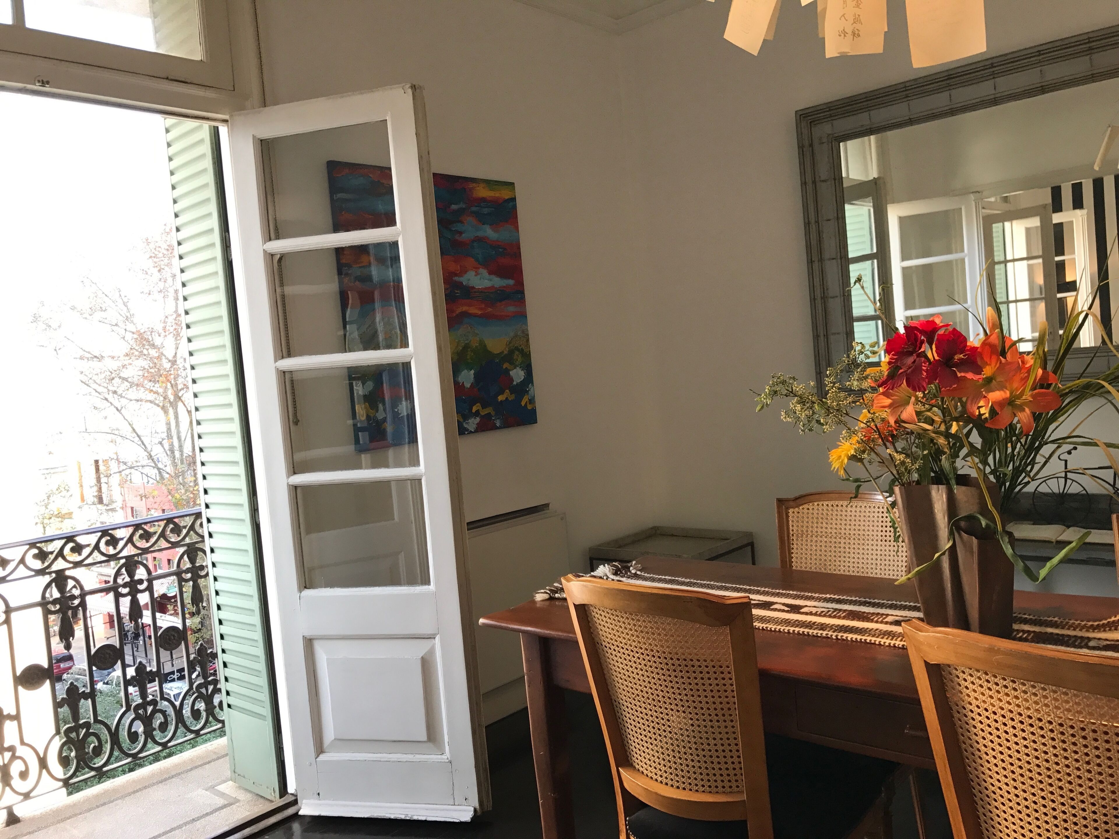 An open door leading to a balcony with a metal railing, a dining table with chairs, a vase with flowers, and two paintings on the wall.